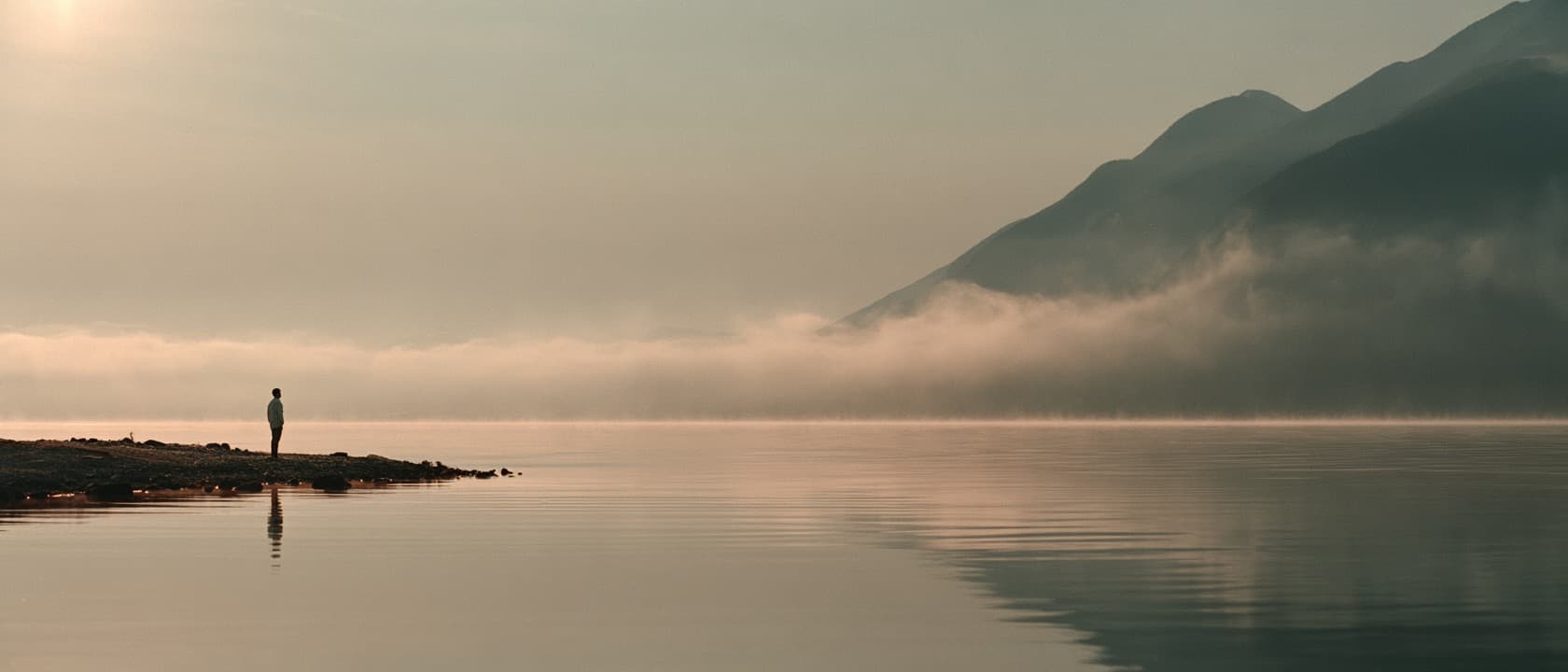 A person standing before a calm lake at dawn, taking in the stillness